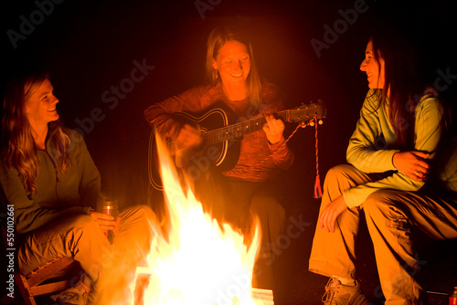 Two women listen, as a third plays the guitar beside a campfire. photo