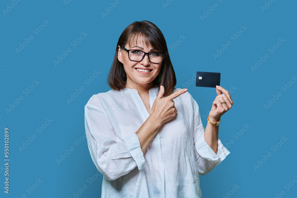 Attractive middle aged smiling woman holding plastic credit card, blue background