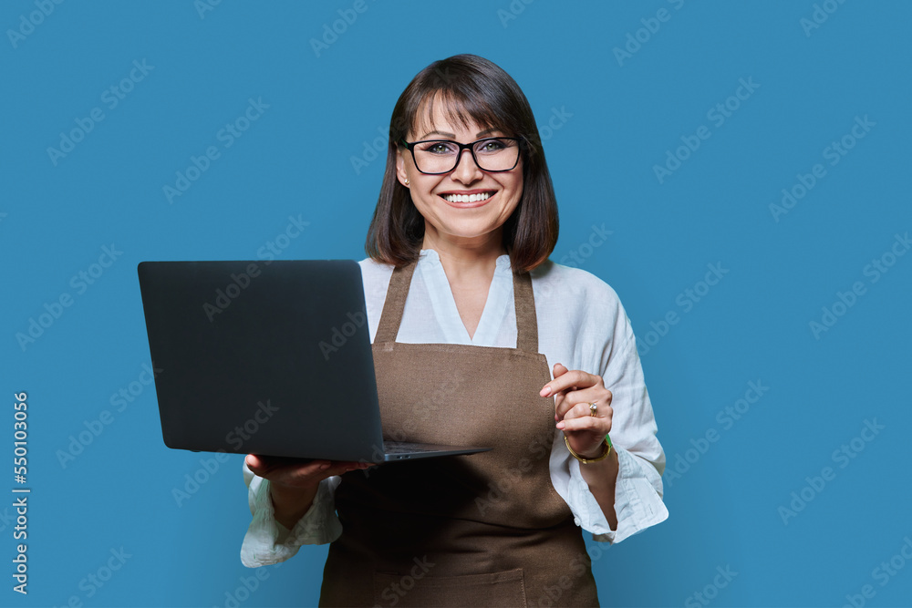 Portrait of middle aged woman in aapron with laptop on blue background. Portrait of middle aged woman in aapron with laptop on blue background.