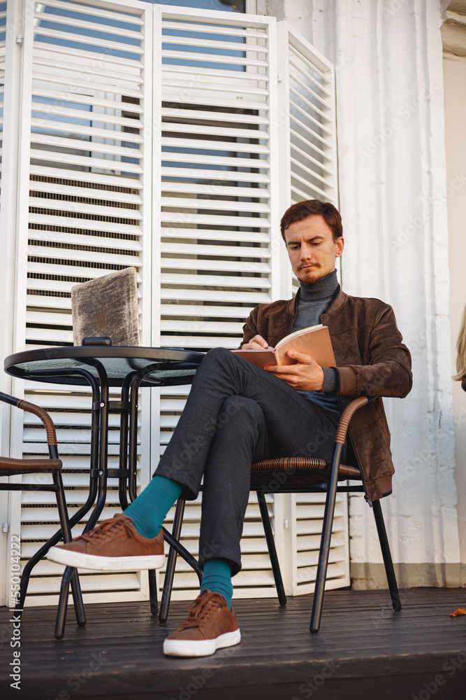 Full length of male freelancer taking notes in planner and working on project remotely in cafe in city street Full length of male freelancer taking notes in planner and working on project remotely in cafe in city street