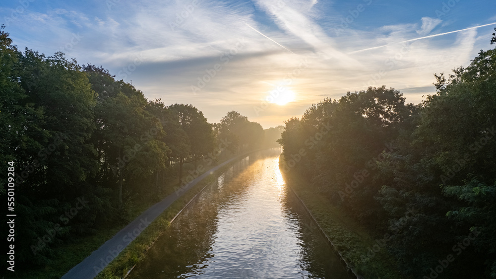 Aerial view, shot by a drone, of the sun rising or setting behind the river and fills the trees with warm evening light, fog rises in the rays of golden light. Clouds and clouds are reflected in the
