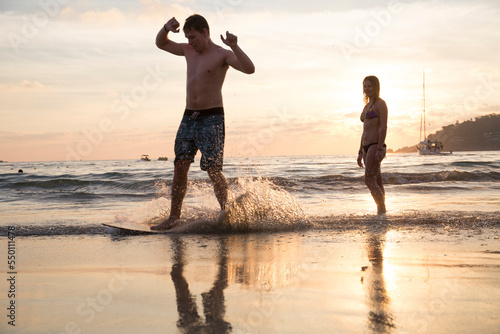 Young man skim boards along beach, girl watches photo