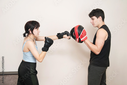 Guy and girl teenagers are training, boy is holding boxing paws and girl is training her kicks for boxing and mixed martial arts, sport concept among teenagers