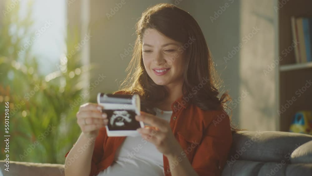 Smiling beautiful pregnant woman watching her ultrasound reports, dreaming about future. Future mother taking preperation for child with baby clothes and shoes Smiling beautiful pregnant woman watching her ultrasound reports, dreaming about future. Future mother taking preperation for child with baby clothes and shoes