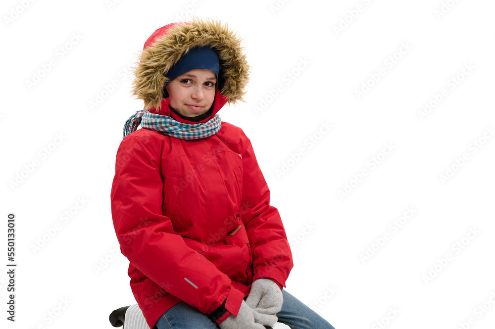 Portrait of multi-ethnic happy teenager traveler boy wearing red parka with fur hood, blue jeans and wool mittens, cutely smiling to camera, isolated over white background with free advertising space