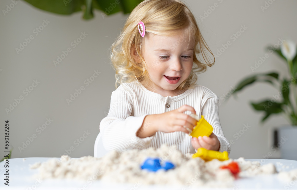 Cute happy caucasian,blonde,curly-haired toddler,baby girl playing with kinetic sand indoors.Preschool kid early development,motor.sensoric skills concept. Cute happy caucasian,blonde,curly-haired toddler,baby girl playing with kinetic sand indoors.Preschool kid early development,motor.sensoric skills concept.