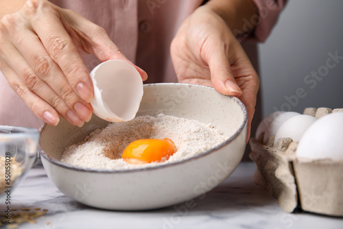 Woman adding egg to bowl with flour at white marble table in kitchen, closeup. Cooking oatmeal dough