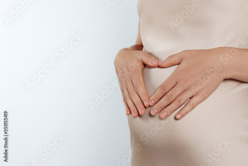 Pregnant woman making heart with her hands on light grey background, closeup. Space for text