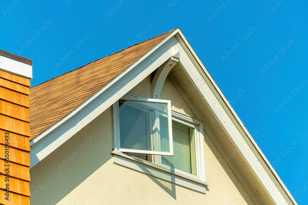 Close up shot with open window in attic of white stucco house with chimney with orange slatted wood with double gable roof on no cloud blue sky background