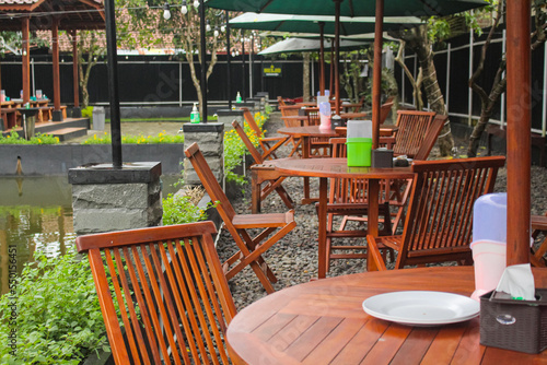 Beautiful and simple outdoor seating area beside the fish pond with industrial wooden tables and chairs and big umbrellas above. Tropical plant decoration for outdoor cafe or restaurant