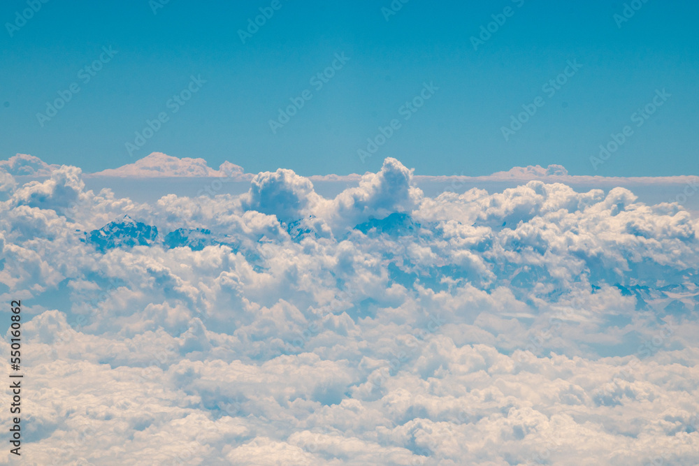 view of a mountain range covered with clouds flying in the blue sky from an airplane window