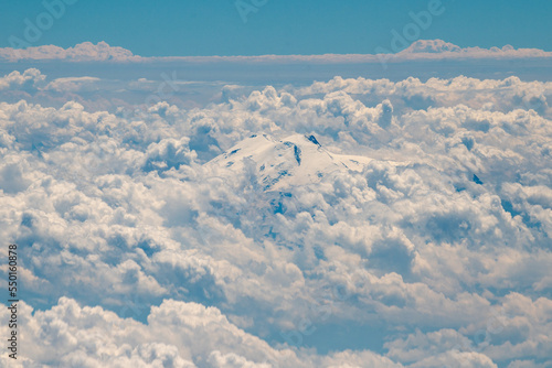 View of the snow-covered peaks of Mount Elbrus, surrounded by white clouds, from the window of an airplane. © fifg