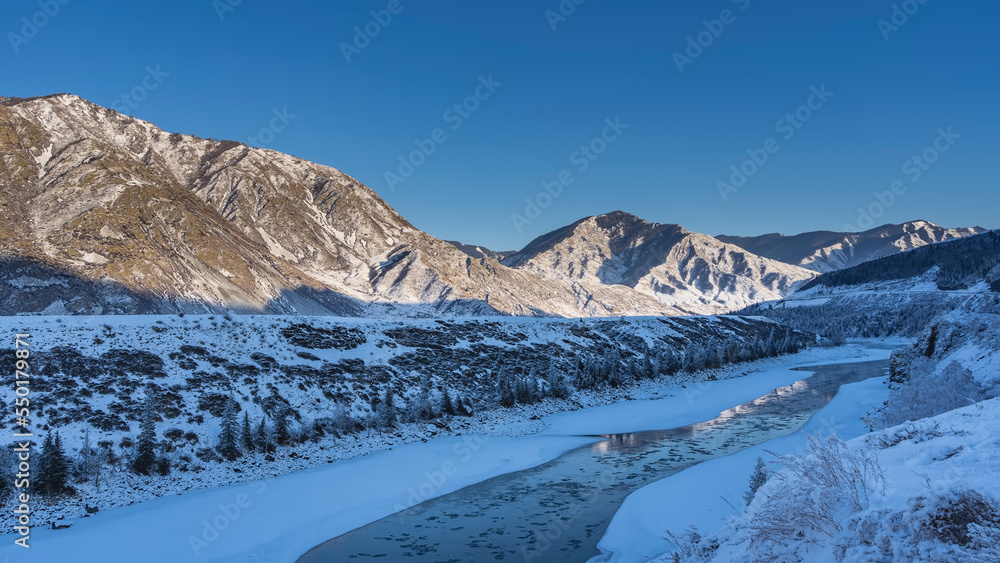 Obraz premium An ice-free river between icy shores. Melted ice and reflection on blue water. Trees and bushes on the snowy shores. The picturesque mountain range is illuminated by the sun against the blue sky.