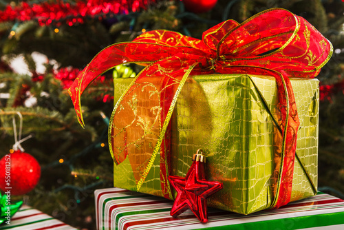 Wrapped Christmas present in gold wrapping paper with a large bow under a decorated Christmas tree
