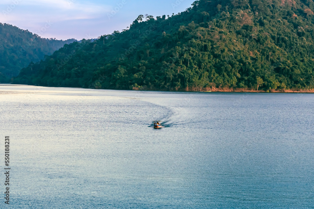 VIew of long-tailed boat floating on blue river in mountain with green forest. River in Khun Dan Prakan Chon Dam in Thailand. Landscape. Tourists on a boat to enjoy scenery in evening. Tourism concept