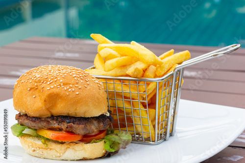 Chicken burger with stainless steel basket of french fries on white plate by swimming pool