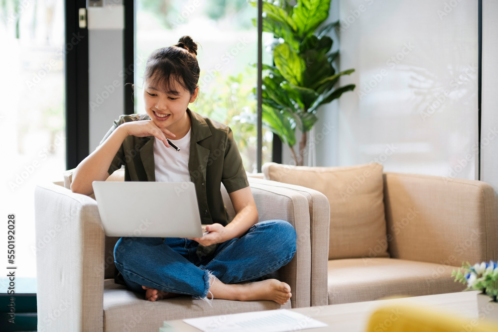 Young asian woman using laptop while leaning back on couch at home.. Young asian woman using laptop while leaning back on couch at home..