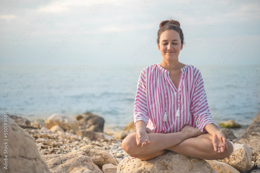 Happy relaxed woman meditation in lotus position practicing yoga sitting on stone beach sea waves