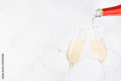 Festive background with flow of pouring fizz champagne from red neck bottle to two glasses on soft light white marble backdrop, copy space, closeup, detail. New Year and celebrate event background. photo