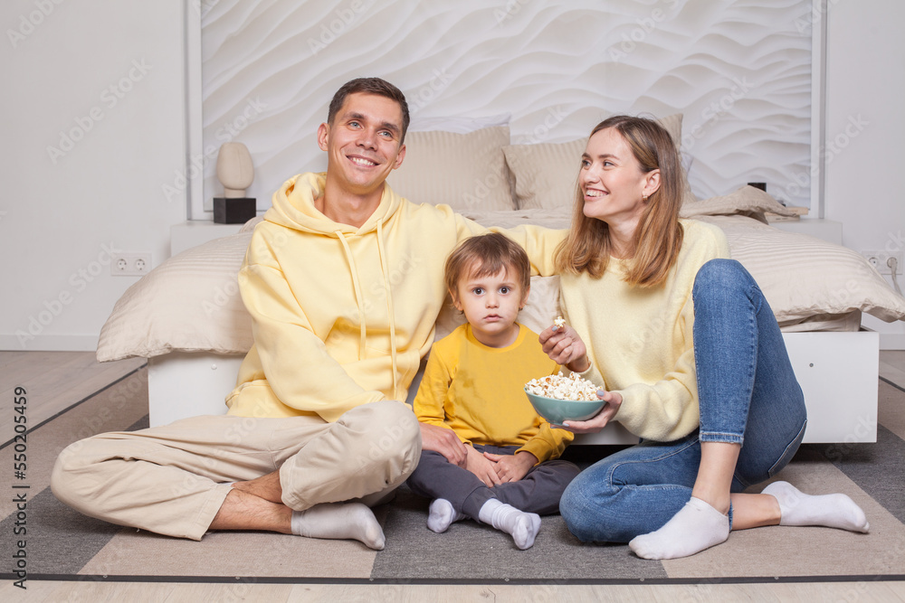 Laughing friendly family, cute parents with child boy son sitting by the bed and eating popcorn Laughing friendly family, cute parents with child boy son sitting by the bed and eating popcorn