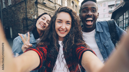 Photos Point of view shot of joyful girls and guys multiethnic group taking selfie holding camera and posing outdoors during enjoyable vacation in beautiful city