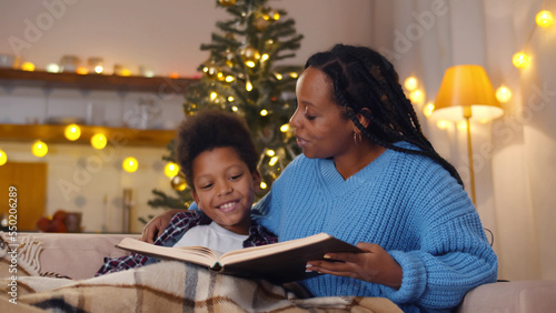 African-American mother and son resting on couch reading book in living room decorated for christmas