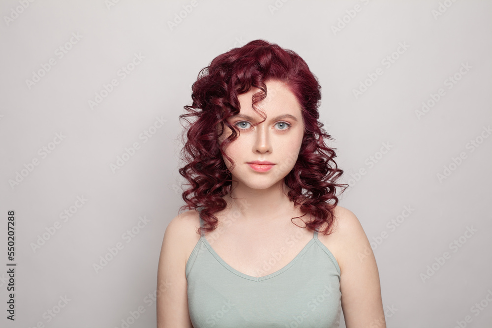 Attractive woman with red curly hair in grey top looking to the camera on white studio wall background Attractive woman with red curly hair in grey top looking to the camera on white studio wall background