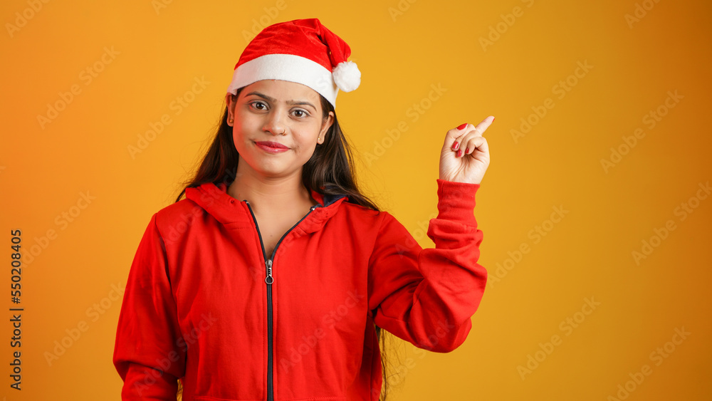 Beautiful young woman pointing empty copy space, girl in Christmas dress and Santa hat, Asian Indian women isolated over colour background Beautiful young woman pointing empty copy space, girl in Christmas dress and Santa hat, Asian Indian women isolated over colour background