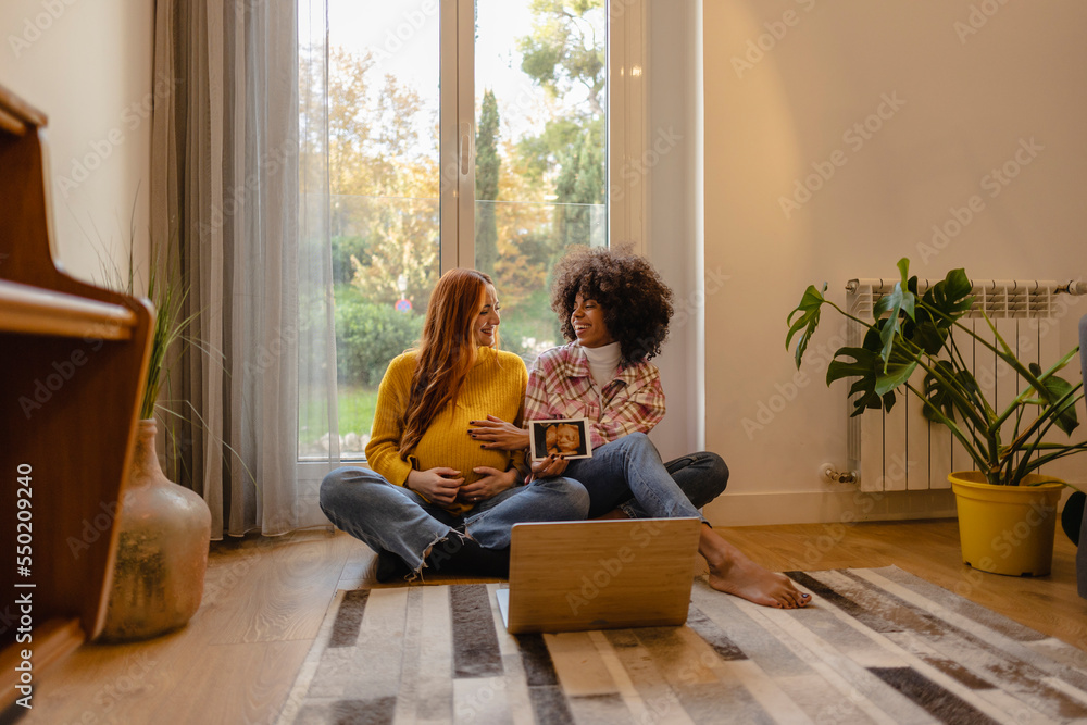 Multiracial couple of two lesbian women sitting on the floor with their baby ultrasound making a video call. Pregnant young redheaded woman