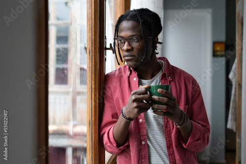 Lonely sad African man looking out window standing indoors suffering due to separation from girlfriend or lack of friends. Unhappy young black guy in casual clothes holding mug with tea or coffee photo