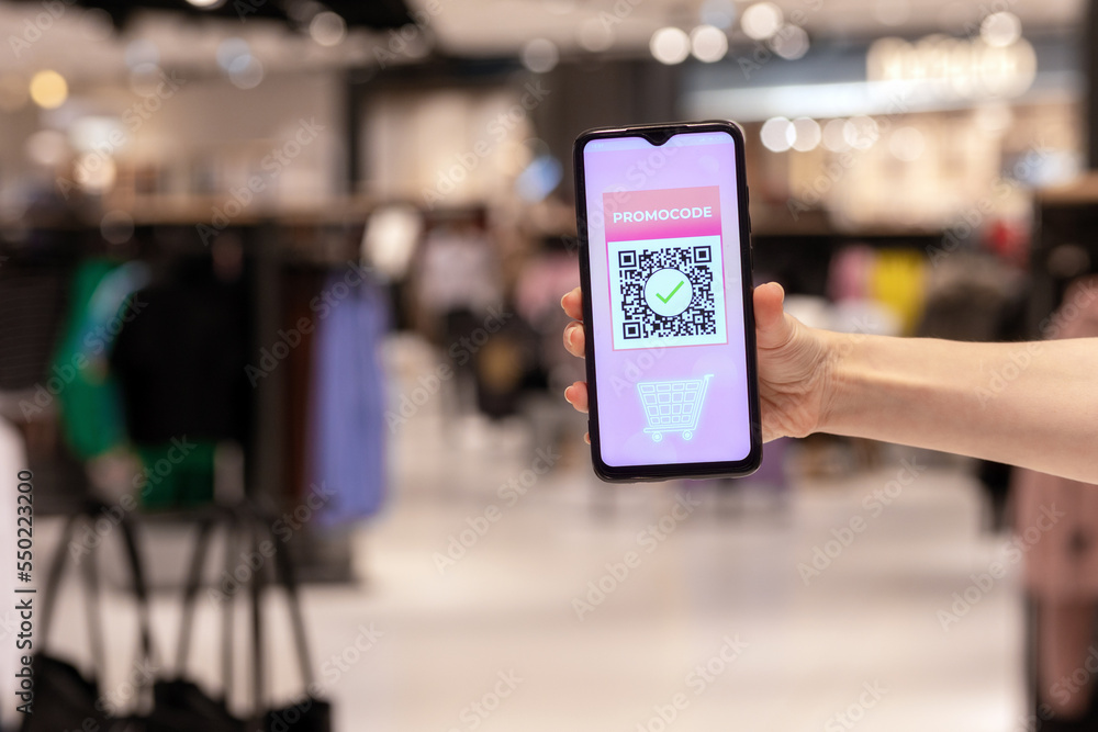 Fototapeta premium Close-up of women's and showing smartphone with purchases in online store. Scan qr code for discount coupon. Defocused shop at background. Concept of shopping and consumerism