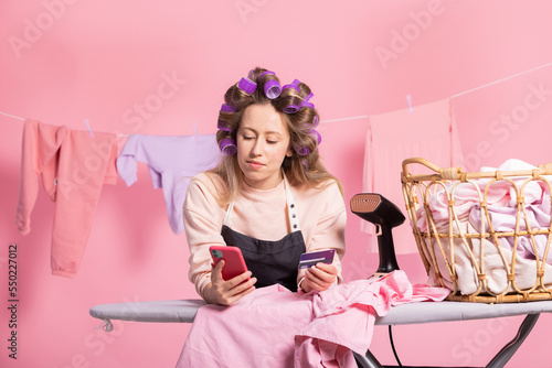 Portrait on pink studio background of housewife in rollers in hair cleaning laundry hanging clothes on string. Woman leans against ironing board and buys groceries over phone while paying with card.