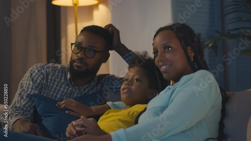 Cheerful African-American family with kid resting on couch talking and watching tv in evening