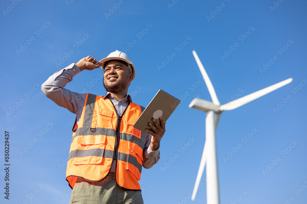 Engineer India man working with tablet at windmill farm Generating electricity clean energy. Wind turbine farm generator by alternative green energy. Asian engineer checking control electric power Engineer India man working with tablet at windmill farm Generating electricity clean energy. Wind turbine farm generator by alternative green energy. Asian engineer checking control electric power