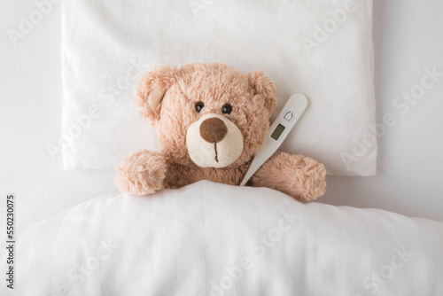 Print op canvas Teddy bear with digital thermometer lying down on pillow and sheet under blanket in white bed