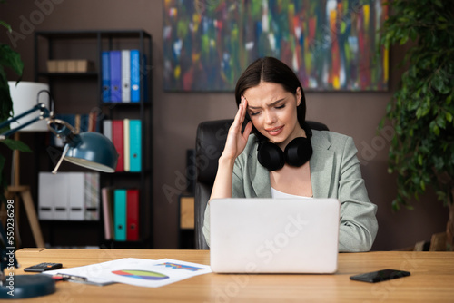 Confused tired worried woman lady girlwearing modern suit and earbuds headphones sitting in front of computer laptop resting relaxing after work process from home.