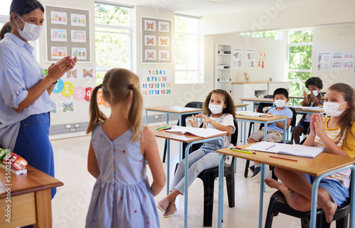 Covid, education and learning with a teacher wearing a mask and clapping for a student after her oral with classmates in class during school. Young girl talking or sharing her answer in a classroom photo