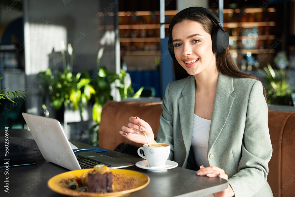 Modern attractive young woman at restaurant sitting with digital devices laptop and earphones looking at camera sniling eating dessert and drinking cofffee during break after work. Working concept.