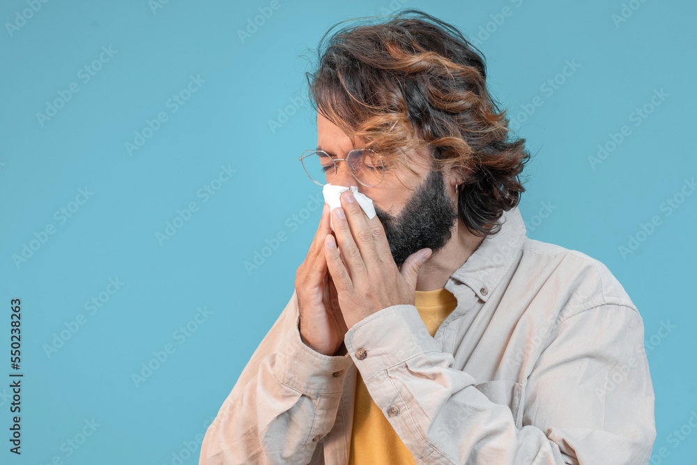 Unhealthy sick man flu sneezing loudly in tissue, feeling unwell with runny nose, fever, caught cold or allergy symptom. Isolated on blue background Unhealthy sick man flu sneezing loudly in tissue, feeling unwell with runny nose, fever, caught cold or allergy symptom. Isolated on blue background