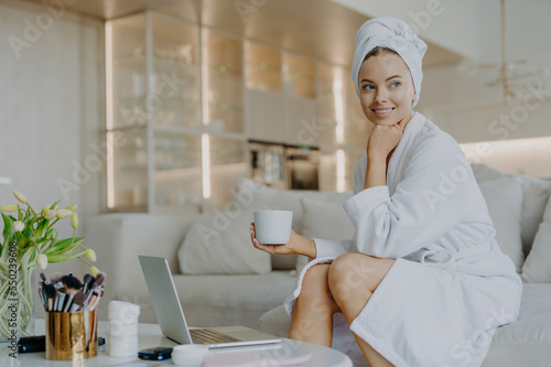 Cheerful refreshed woman with healthy skin wears bathrobe after taking shower drinks coffee and looks thoughtfully aside sits on comfortable sofa works on laptop computer uses cosmetic products