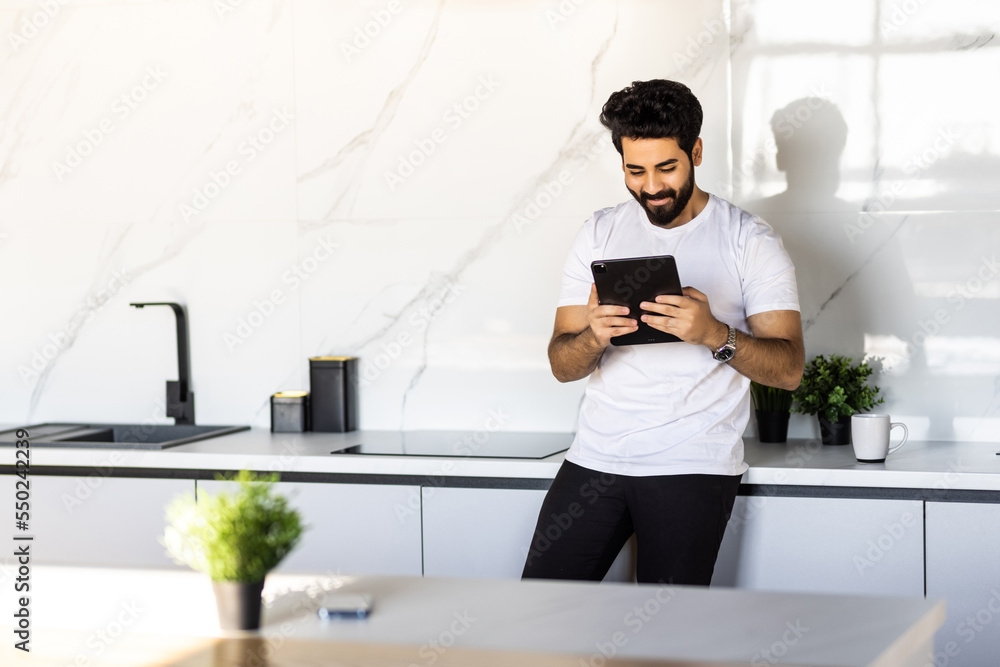 Leisure time with device. Happy arab man reading e-book on digital tablet or surfing internet while sitting in kitchen and enjoying morning coffee at home