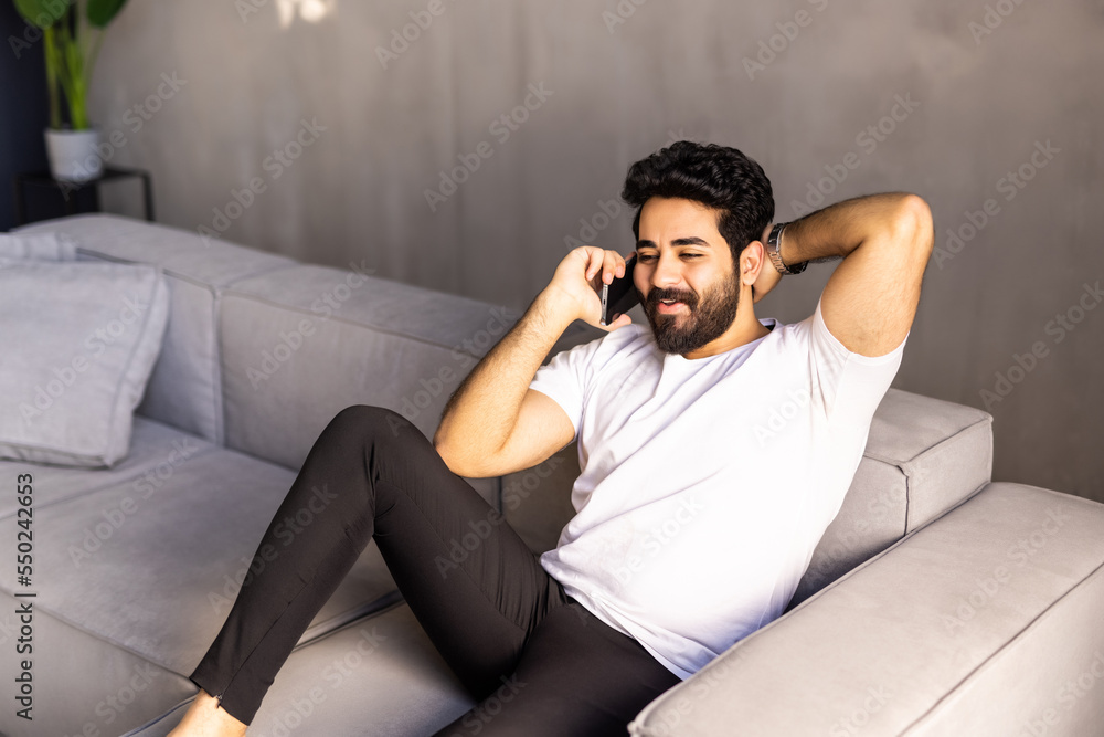 Fototapeta premium Cheerful arab man with long hair and glasses relaxing on couch at home, having phone talk with friends, looking at copy space and smiling, wearing casual outfit, resting at weekend