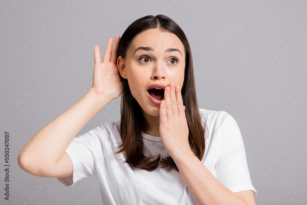 Young curious inquisitive woman girl lady standing on grey background in studio isolated holding hand near ears trying to overhear new information gossip about somebody or discounts. Young curious inquisitive woman girl lady standing on grey background in studio isolated holding hand near ears trying to overhear new information gossip about somebody or discounts.
