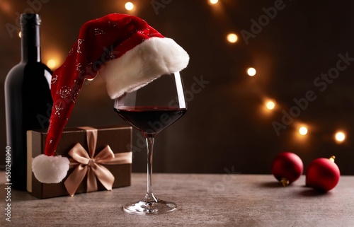 Glass of red wine with Santa Claus hat, bronze gift box and Christmas decorations on table. Christmas lights in the black background.