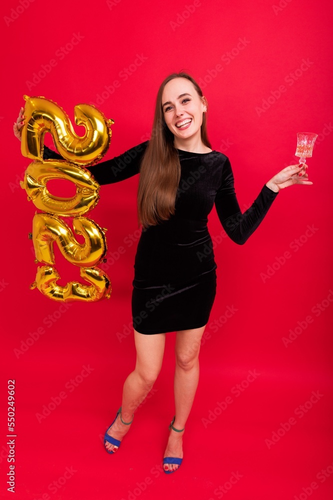 A young woman in a black dress and heels drinks champagne from a glass and holds the figures of the new year 2023 on a red background A young woman in a black dress and heels drinks champagne from a glass and holds the figures of the new year 2023 on a red background