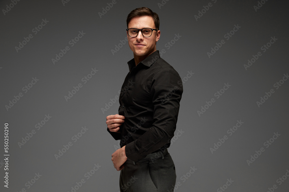 Smiling confident young man in black business classic style shirt and grey trousers posing isolated over grey background. Fashion look of businessman. Office style Smiling confident young man in black business classic style shirt and grey trousers posing isolated over grey background. Fashion look of businessman. Office style