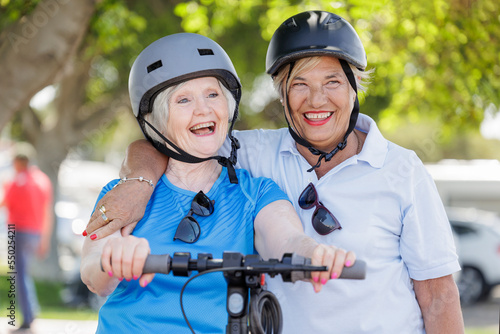 coppia di anziane con caschetto di protezione si abbraccia felice in una pista ciclabile con vicino un monopattino elettrico 