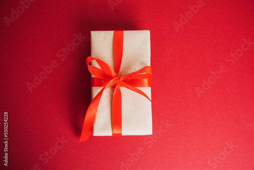 A gift wrapped in brown paper with a red ribbon on a red background. Christmas gift, flat lay