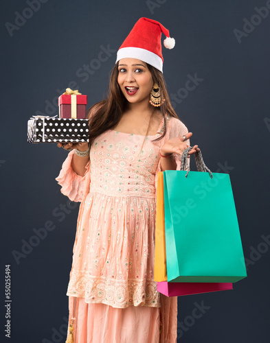 Happy excited young woman in a Santa Claus hat holding shopping bags with gift boxes over gray background, Christmas concept.