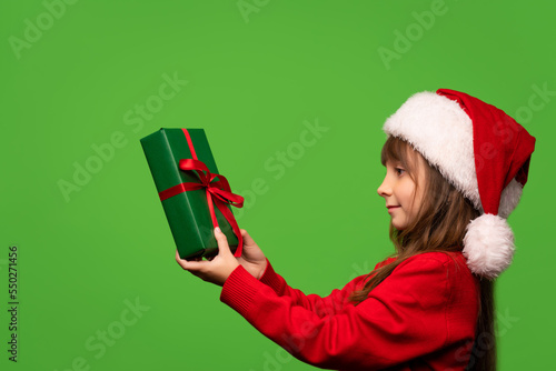 Cute little Santa girl in a hat holds a New Year's gift in her hands and looks at him. Happy New Year. Isolated on green background